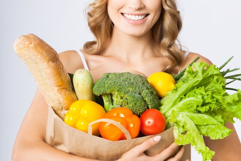 Portrait of happy smiling young beautiful woman holding grocery shopping bag with healthy vegetarian raw food, over gray background